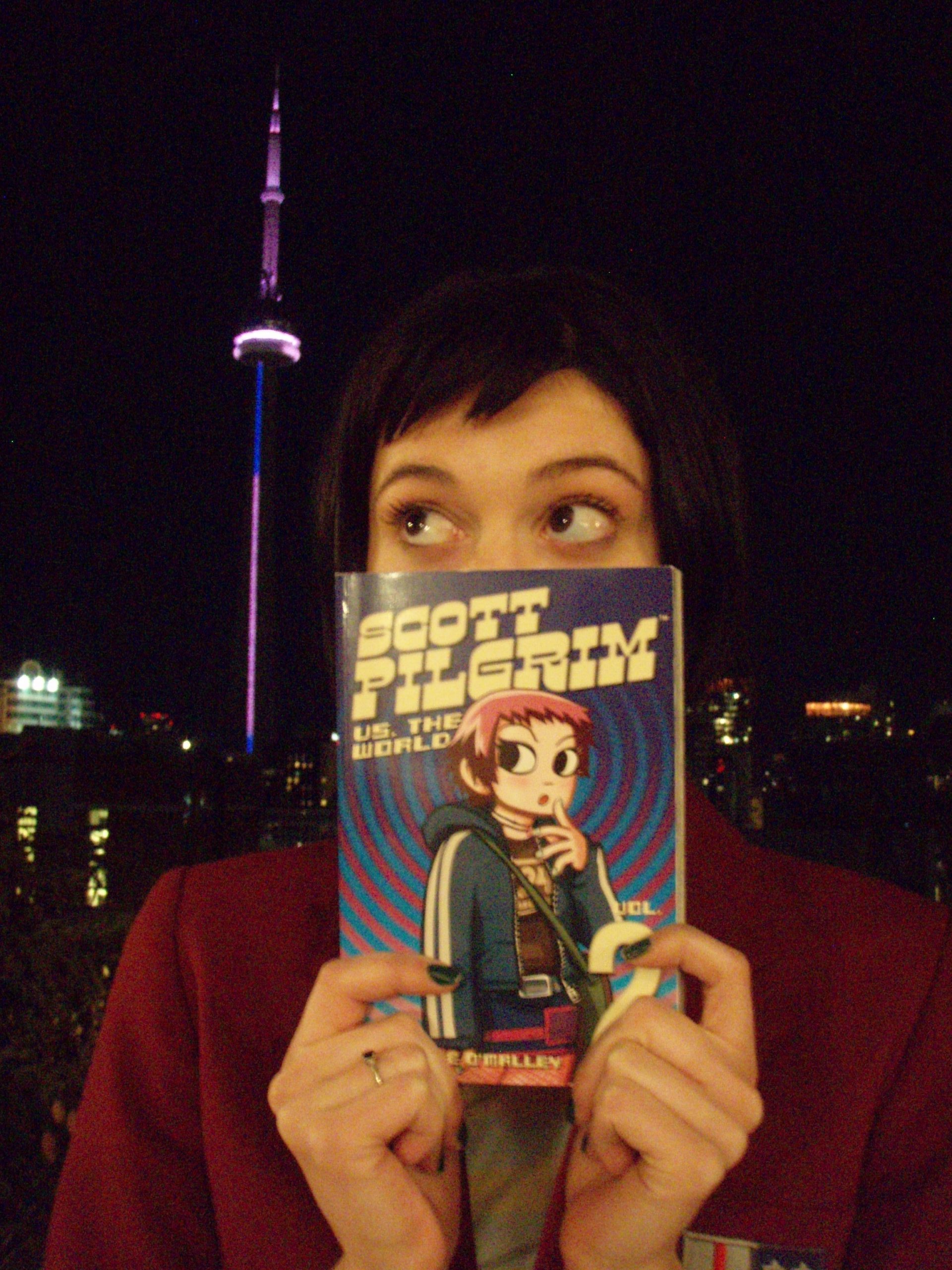 Mary Elizabeth Winstead in Toronto holding a book in front of half of her face. The book is Scott Pilgrim Vol 2 and has an illustration of Ramona Flowers on the cover.