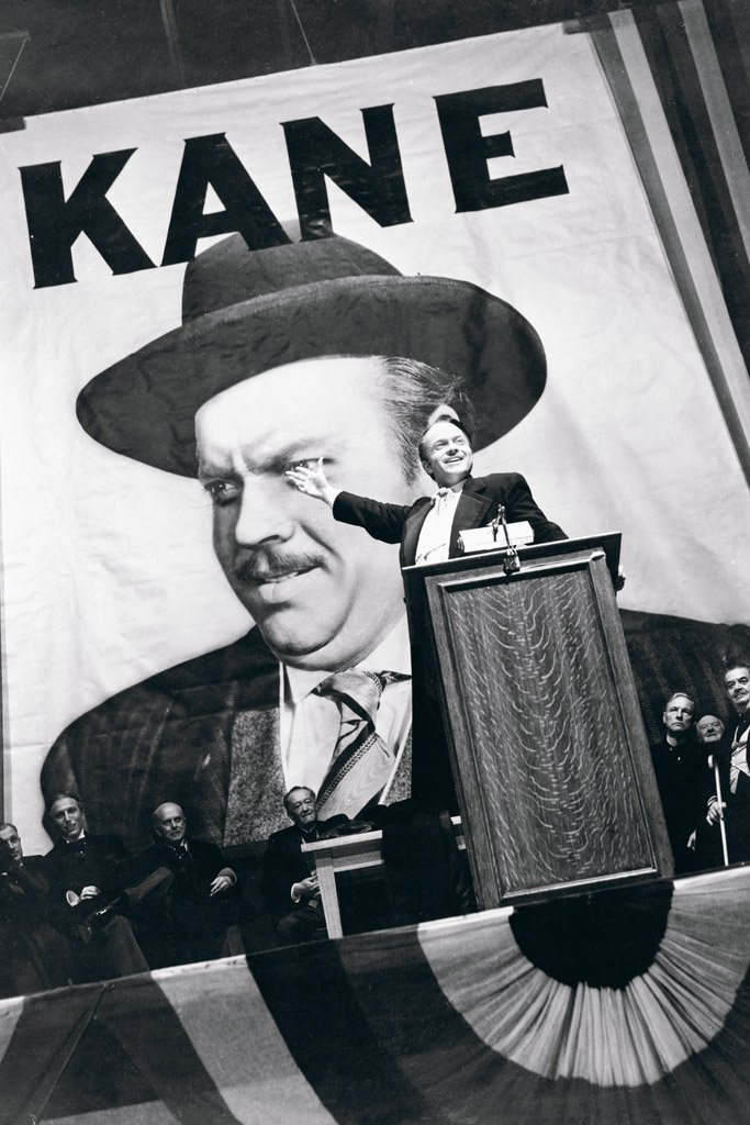 Kane running for governor standing on a stage point at a huge picture of himself behind him